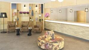 Ein Empfangsbereich mit einem Holzbüro, drei gepolsterten Stühlen, einem runden Couchtisch und Regalen mit Büchern und Blumen.