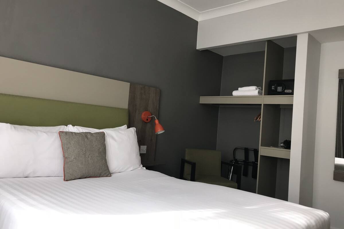 A hotel room with a large bed, a nightstand, a red lamp, a chair, and a shelf with towels and a radio.