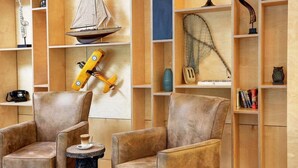 Ein Zimmer mit zwei braunen Sesseln, einem kleinen runden Tisch und Holzregalen mit verschiedenen Gegenständen, darunter ein Modellsegelschiff, ein Spielzeugflugzeug und ein Fischernetz.