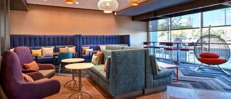 A modern lounge area with a blue sofa, purple armchair, and a hanging chair by a large window with a view of parked cars.