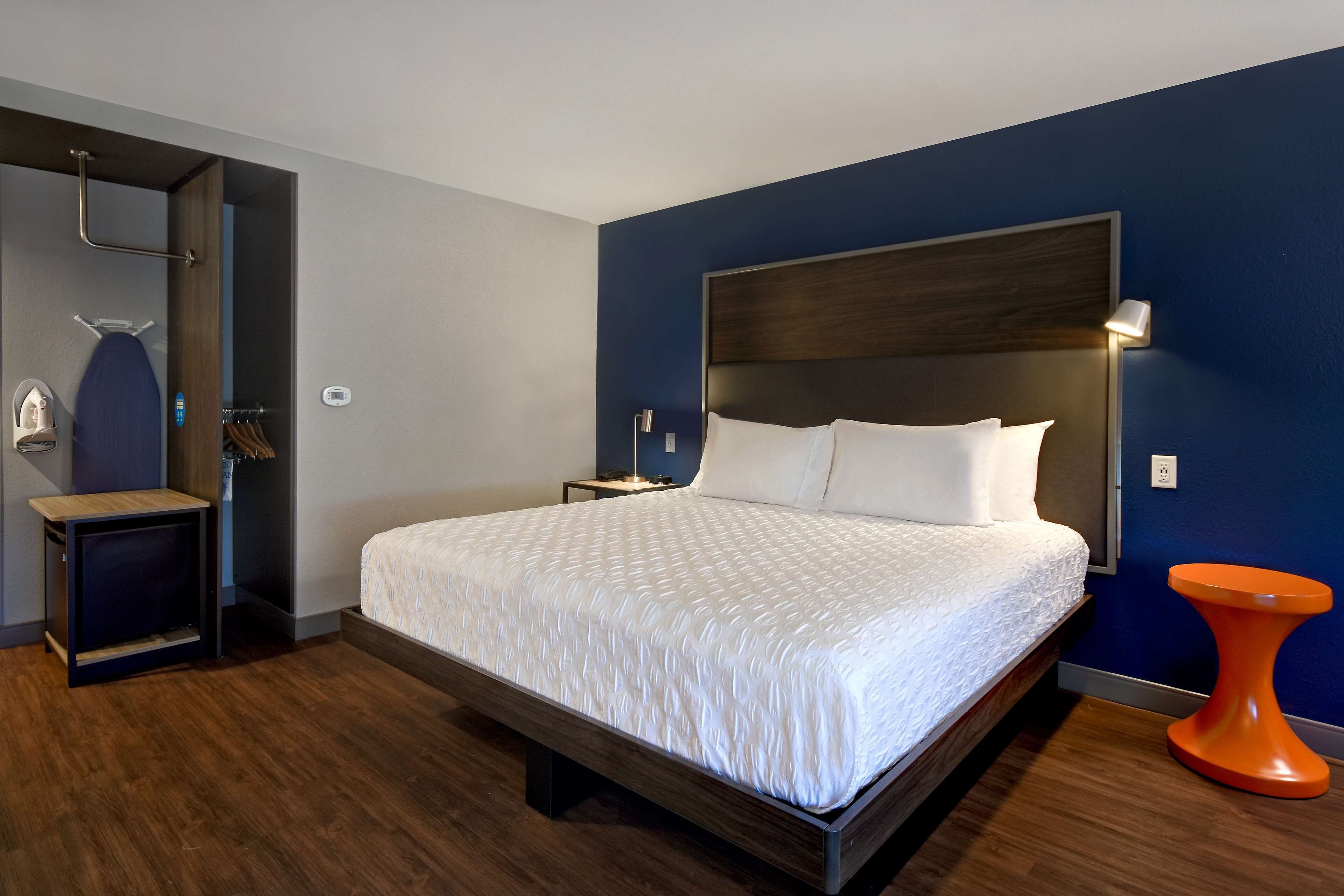 A modern hotel room with a large bed, a bedside table, a nightstand, a wall-mounted lamp, and a unique orange stool.
