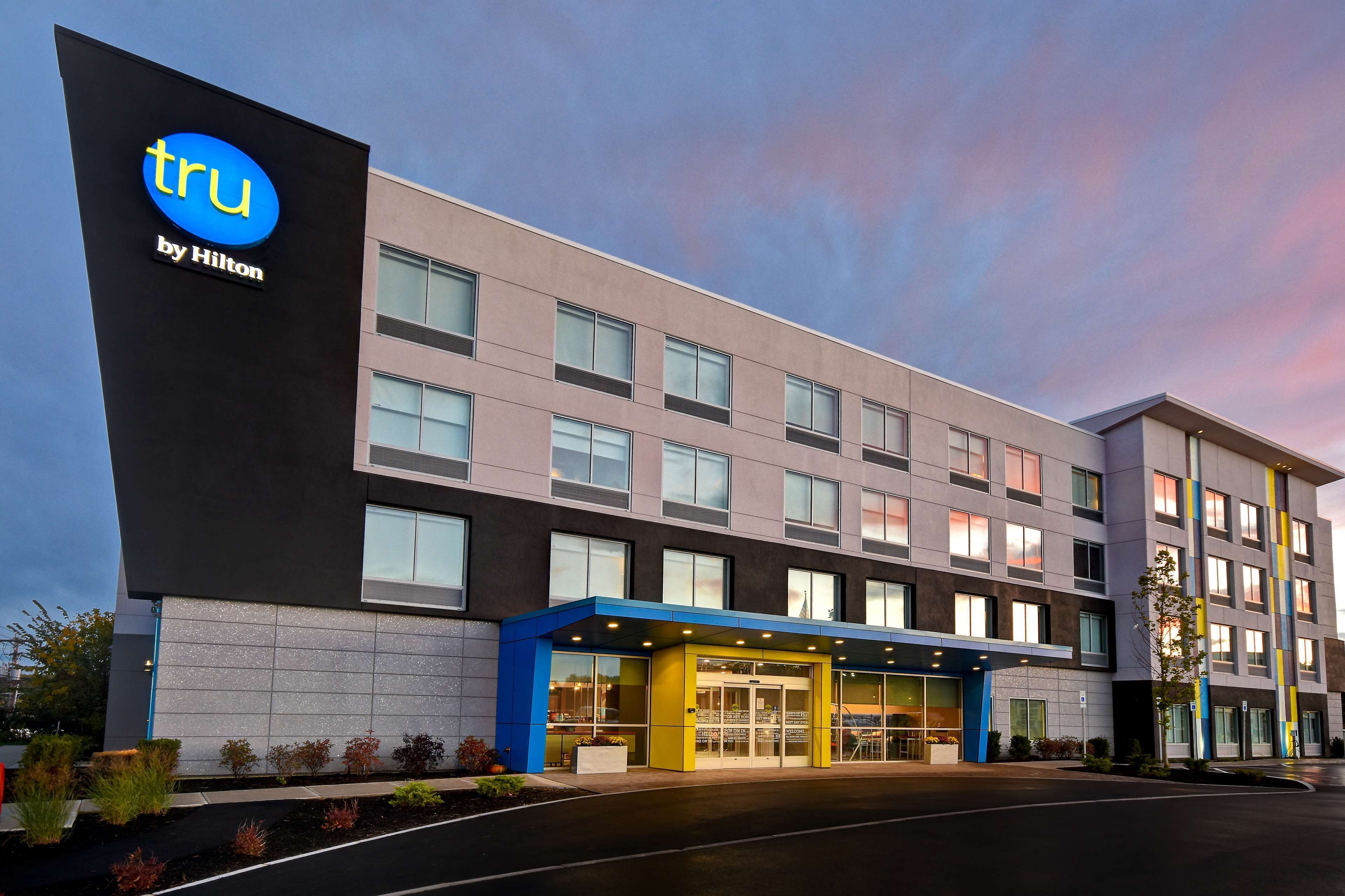 A modern hotel building with a prominent 'tru by Hilton' sign, large windows, and a covered entrance area.