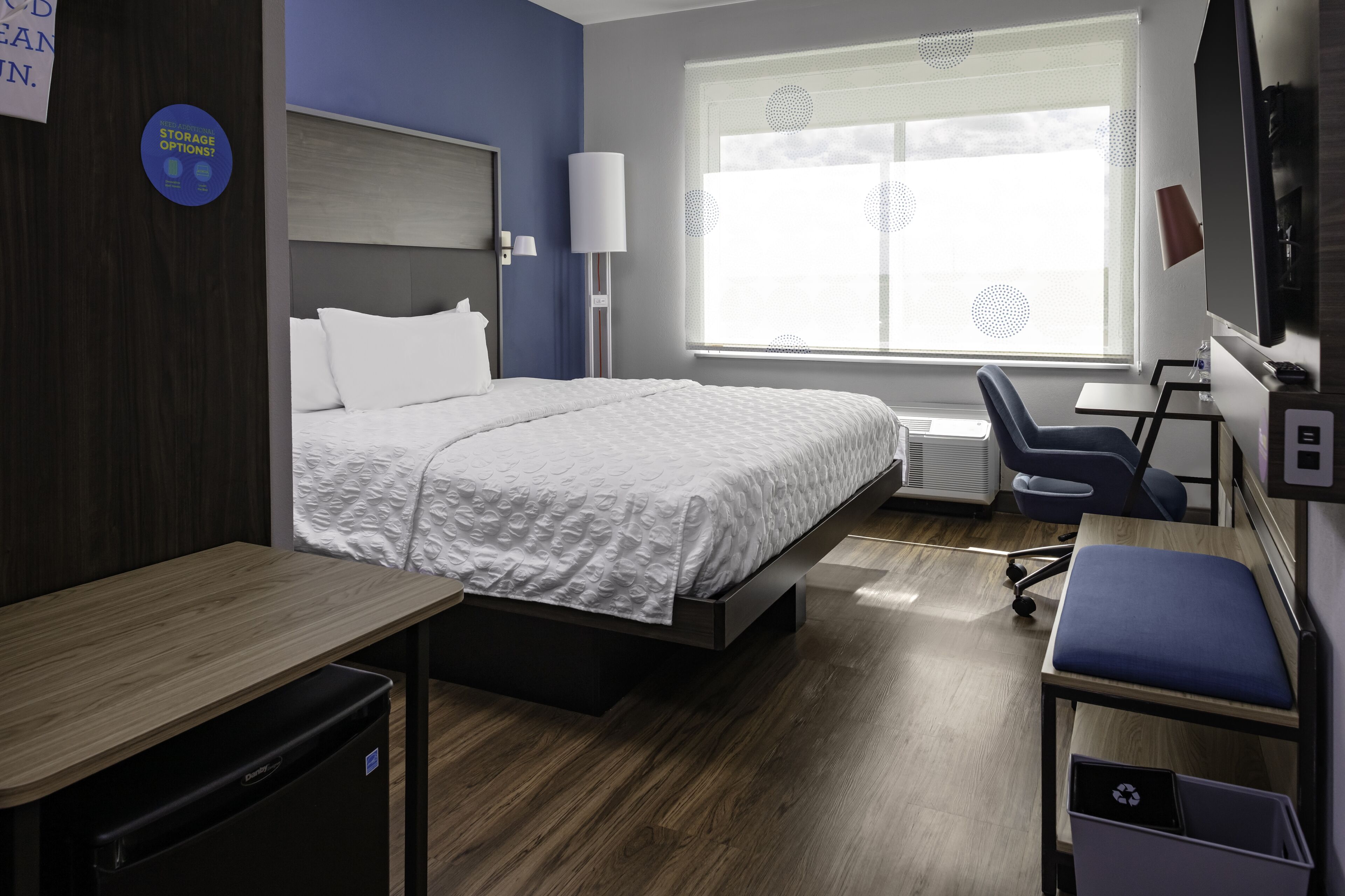 A hotel room with a bed, desk, chair, and a window with blinds.