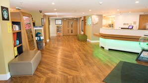 A reception area with a wooden floor, a reception desk, and a green cushioned bench. - Holiday Inn Slough - Windsor by IHG (Slough)