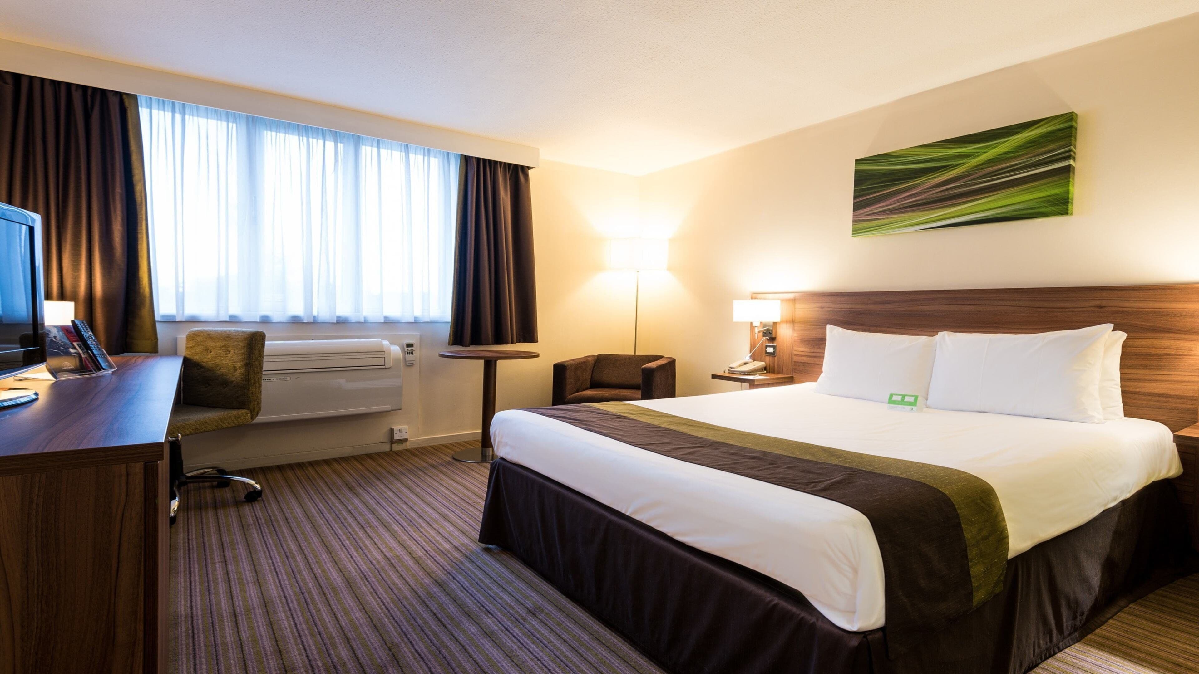 Una habitación de hotel con una cama, un escritorio con una computadora, una silla, una lámpara y un cuadro en la pared.