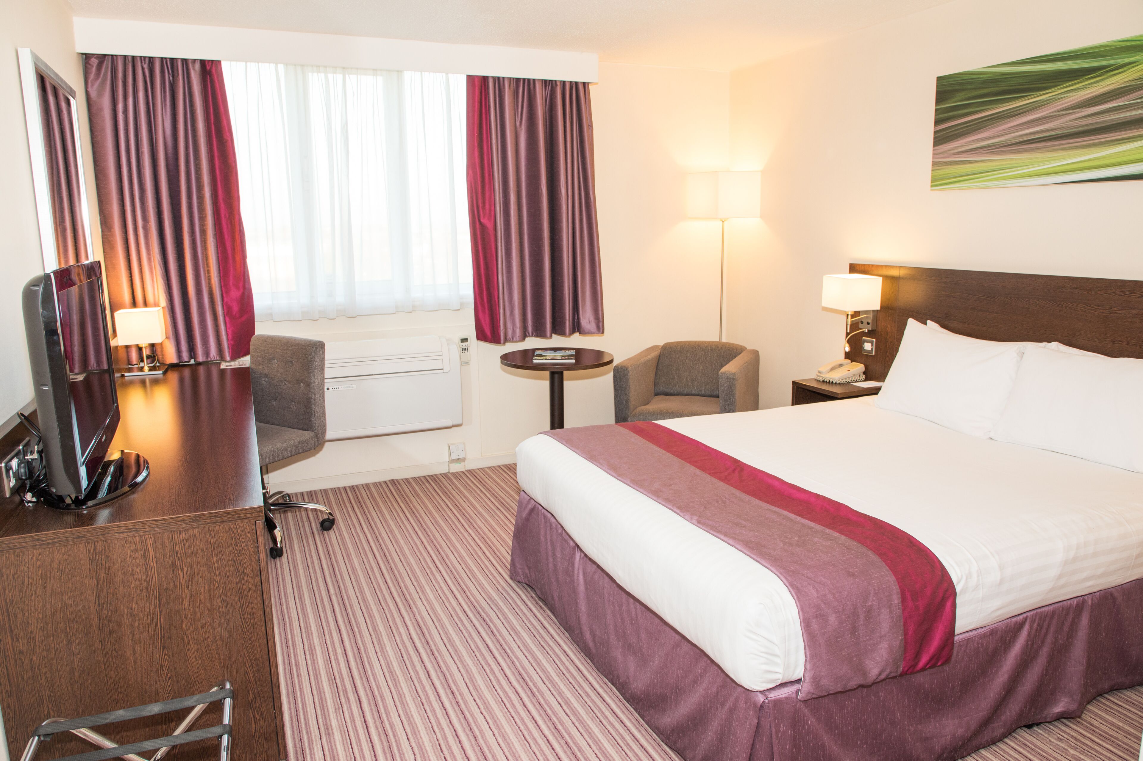 A hotel room with a large bed, a desk with a computer, a chair, a small table, and a lamp.