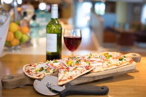 A pizza on a wooden board with a pizza cutter, a bottle of wine, and a glass of red wine on a bar counter.