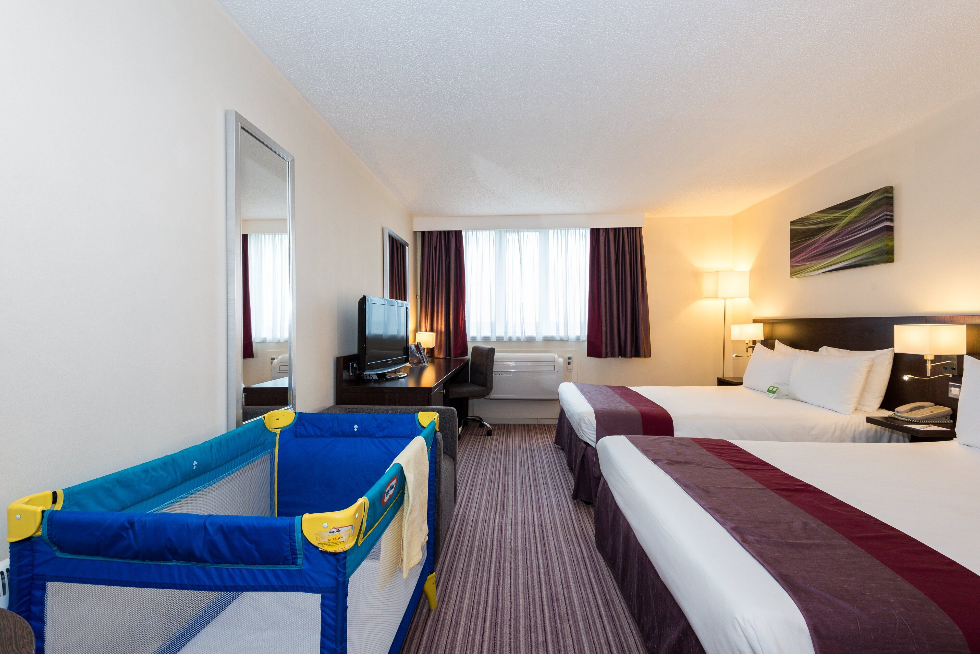 A hotel room with a bed, a desk with a TV, a small playpen, and a window with curtains.