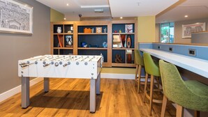 A room with a foosball table, a bar area with stools, and shelves with various items including a LEGO play sign. - Holiday Inn Slough - Windsor by IHG (Slough)