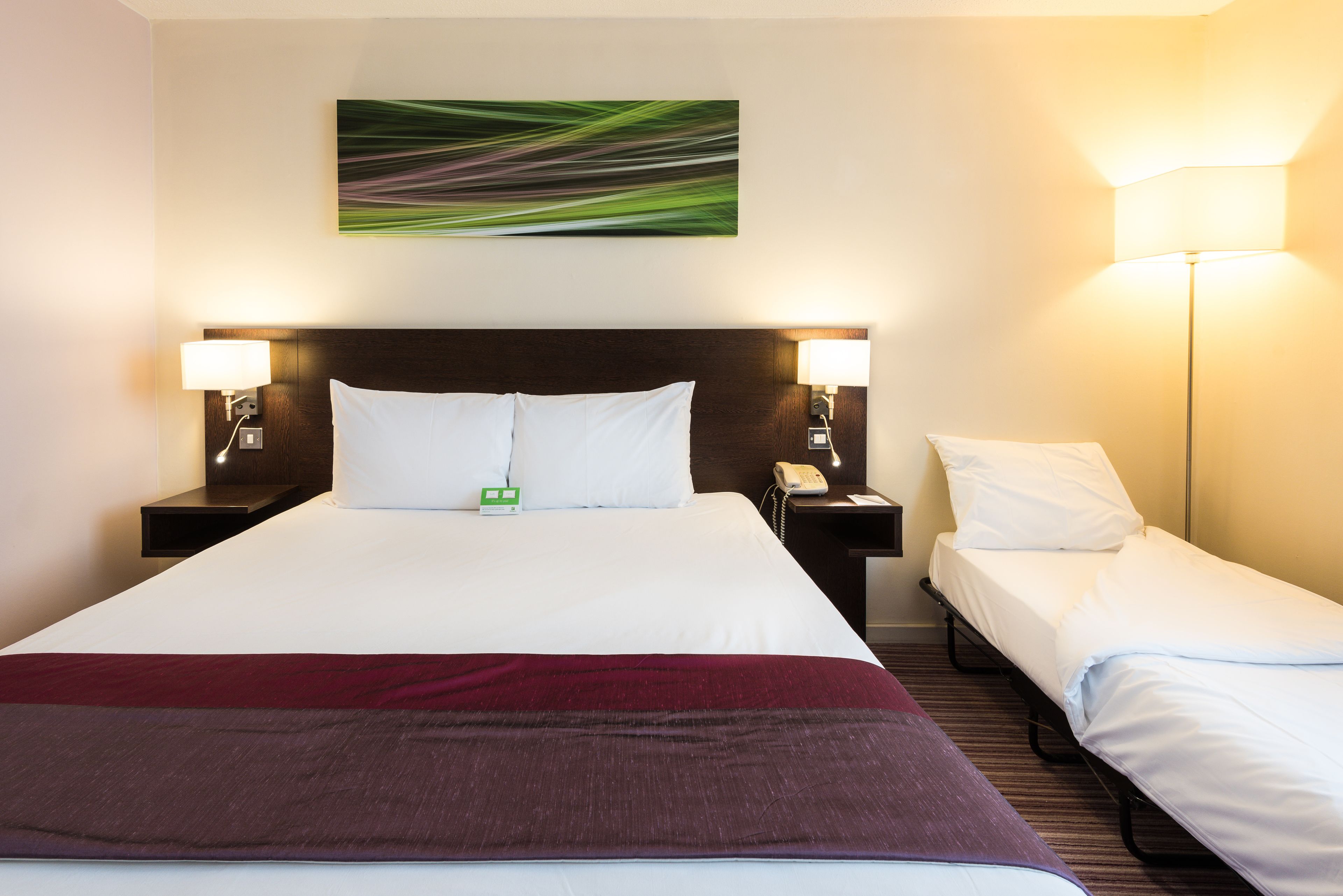 A hotel room with a large bed, two bedside tables, a bedside lamp, a small table with a phone, and a wall-mounted artwork.