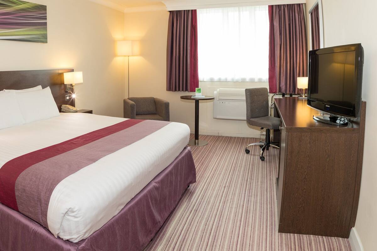 Ein Hotelzimmer mit einem großen Bett, einem Schreibtisch mit Fernseher, einem Sessel, einem kleinen Tisch und einer Lampe.