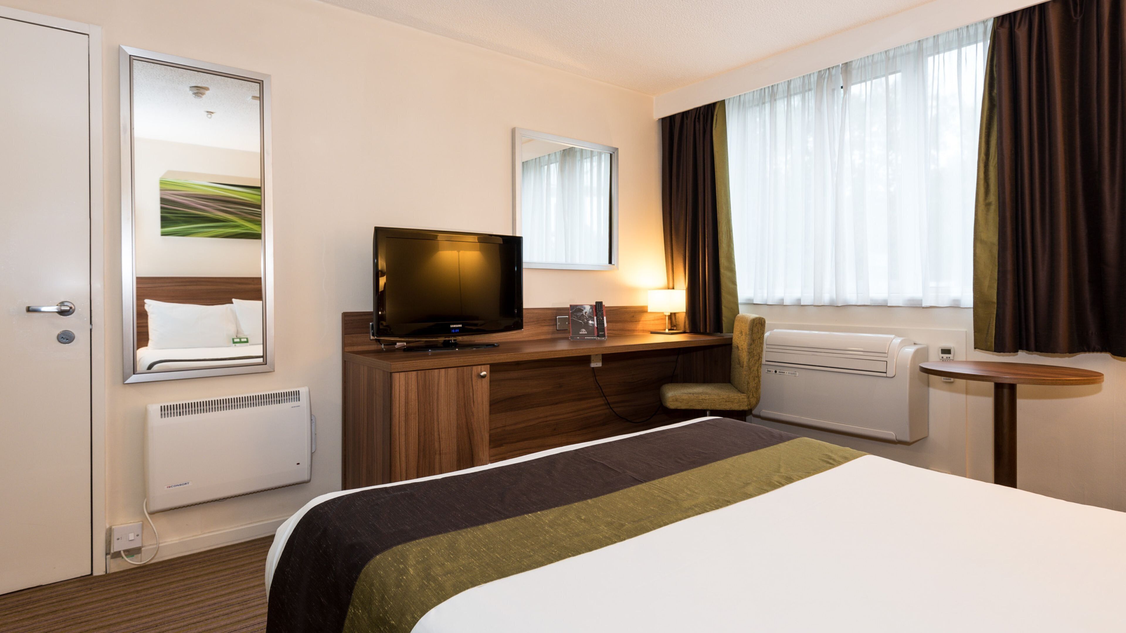 Hotelkamer met een bed, een bureau met een televisie, een stoel, een raam met gordijnen en een radiator.