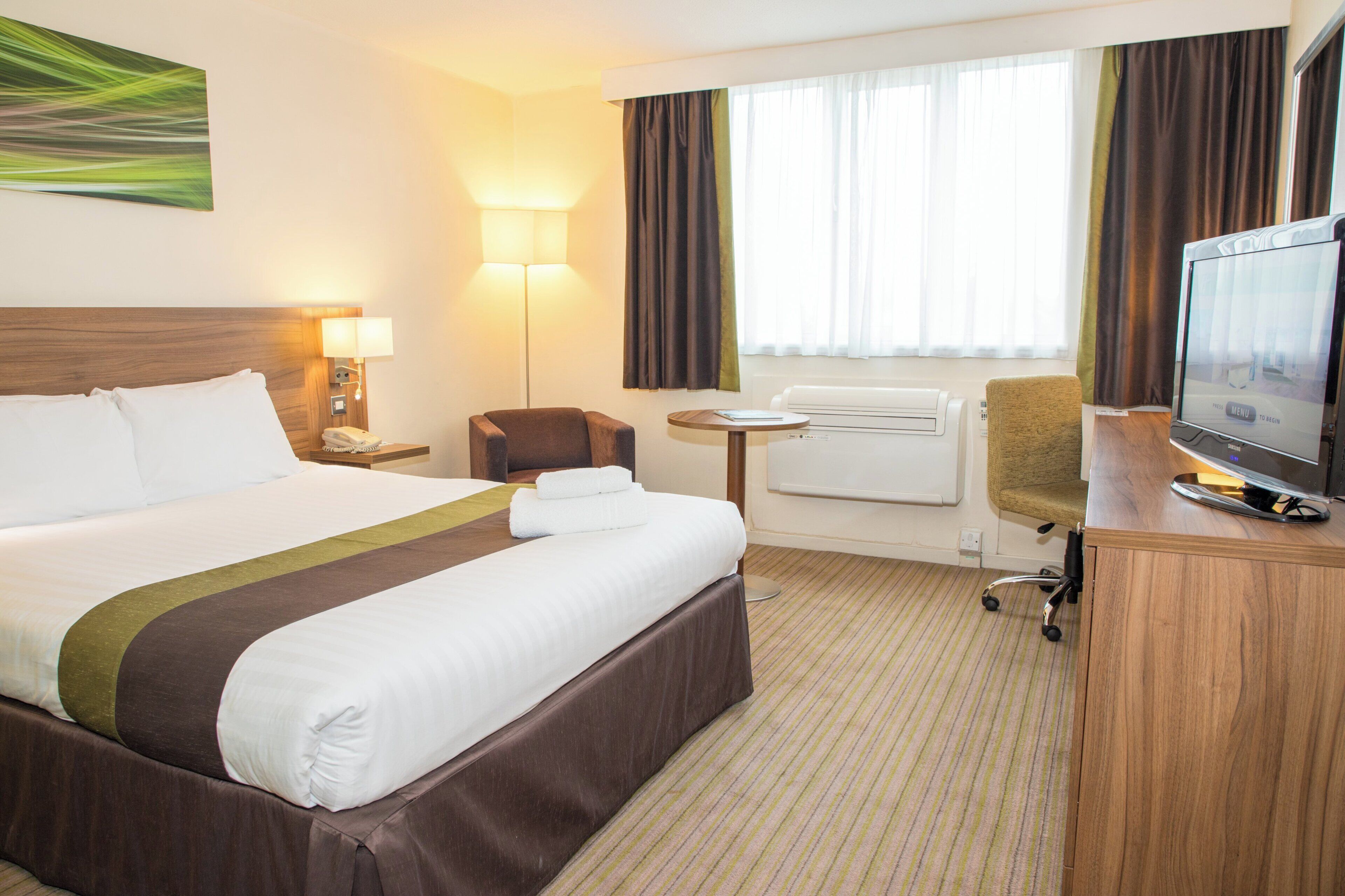 A hotel room with a large bed, a desk with a computer, a chair, a flat-screen TV, a lamp, and a window with curtains.