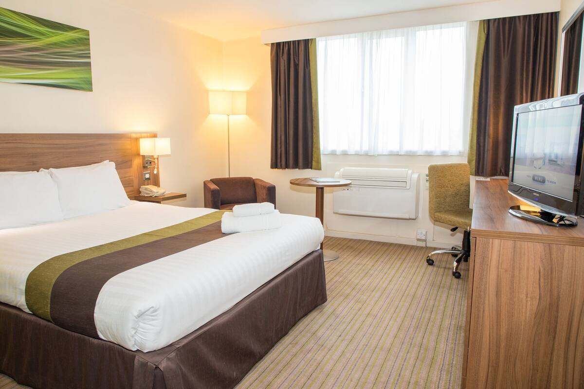 Ein Hotelzimmer mit einem großen Bett, einem Schreibtisch mit Fernseher, einem Sessel, einer Lampe und einem Fenster mit Vorhängen.