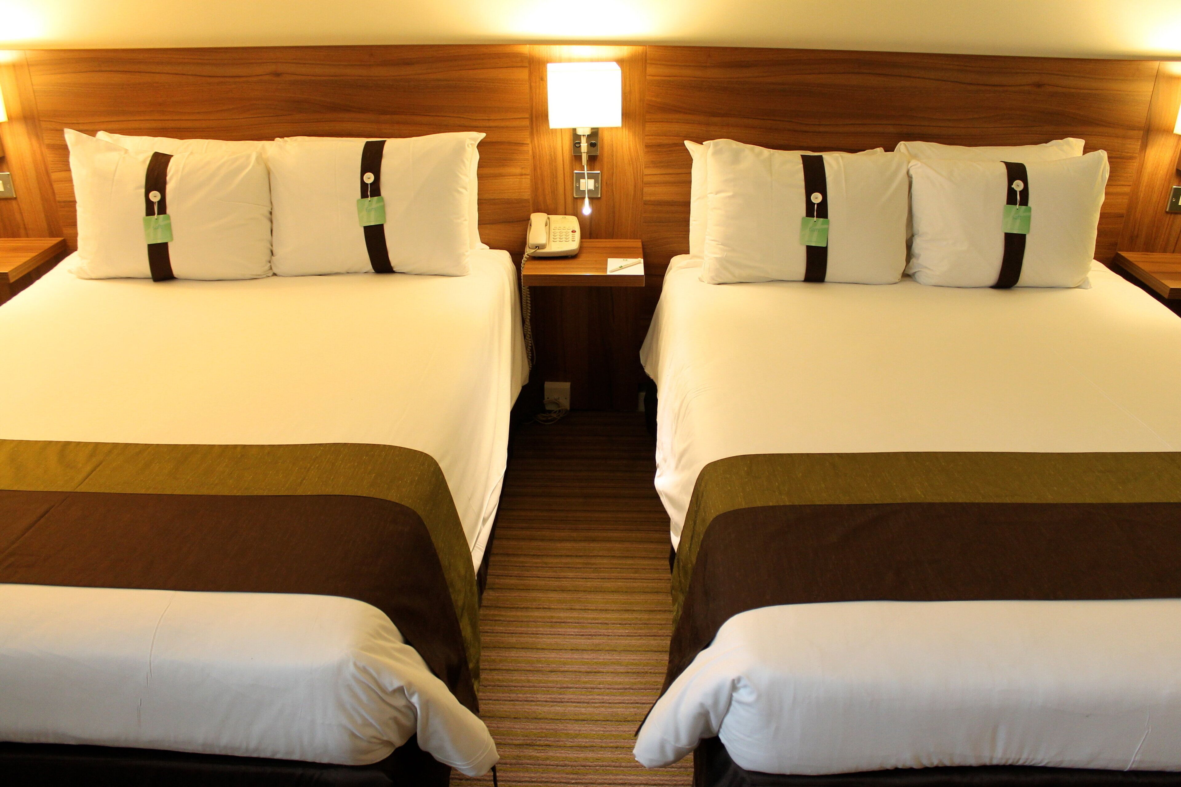 A hotel room with two beds, each with white bedding and brown headboards. There is a bedside table with a lamp and a telephone.