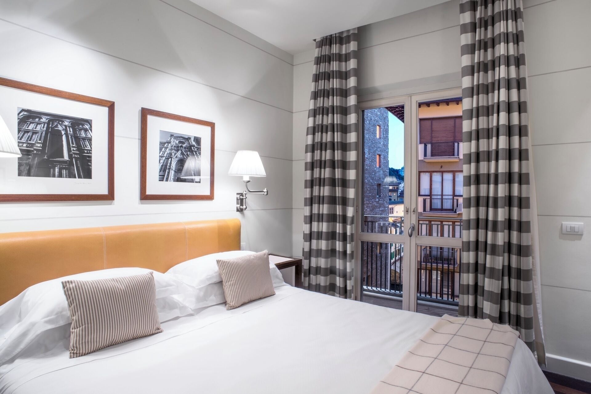 A hotel room with a bed, two framed pictures on the wall, a nightstand with a lamp, and a balcony with striped curtains.