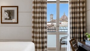 A hotel room with a bed, a chair, a table with a vase, and a view of buildings through a window with striped curtains. - Gallery Hotel Art - Lungarno Collection (Florence)