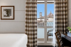 Une chambre d’hôtel avec un lit, une chaise, une table avec un vase, et une vue sur les bâtiments à travers une fenêtre ornée de rideaux à rayures.