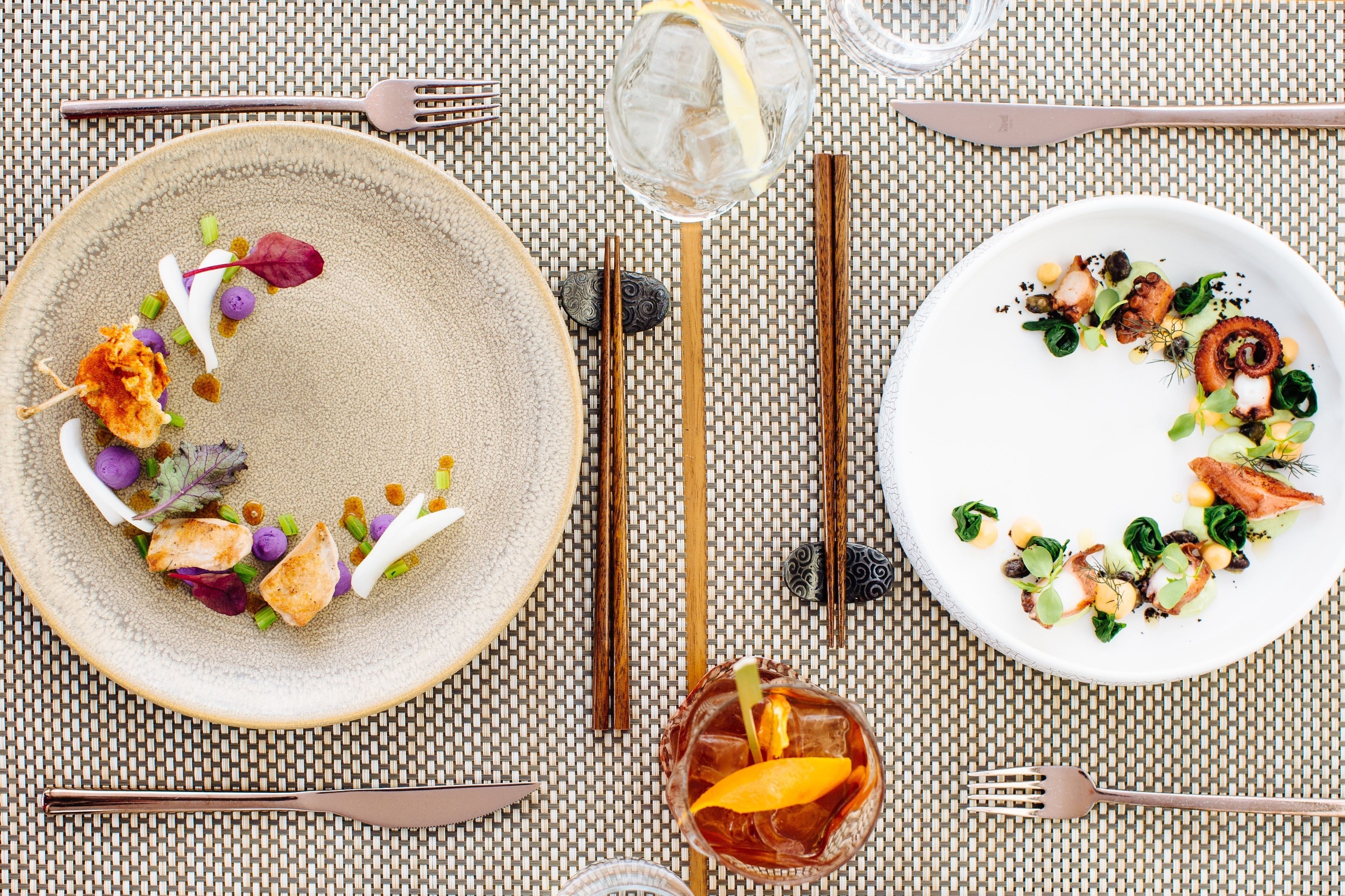 A dining table set with plates of food, chopsticks, and a glass of iced drink.