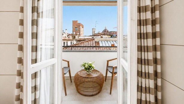 A balcony with a view of buildings, a wicker chair, and a small table with a vase. - Gallery Hotel Art - Lungarno Collection (Florence)