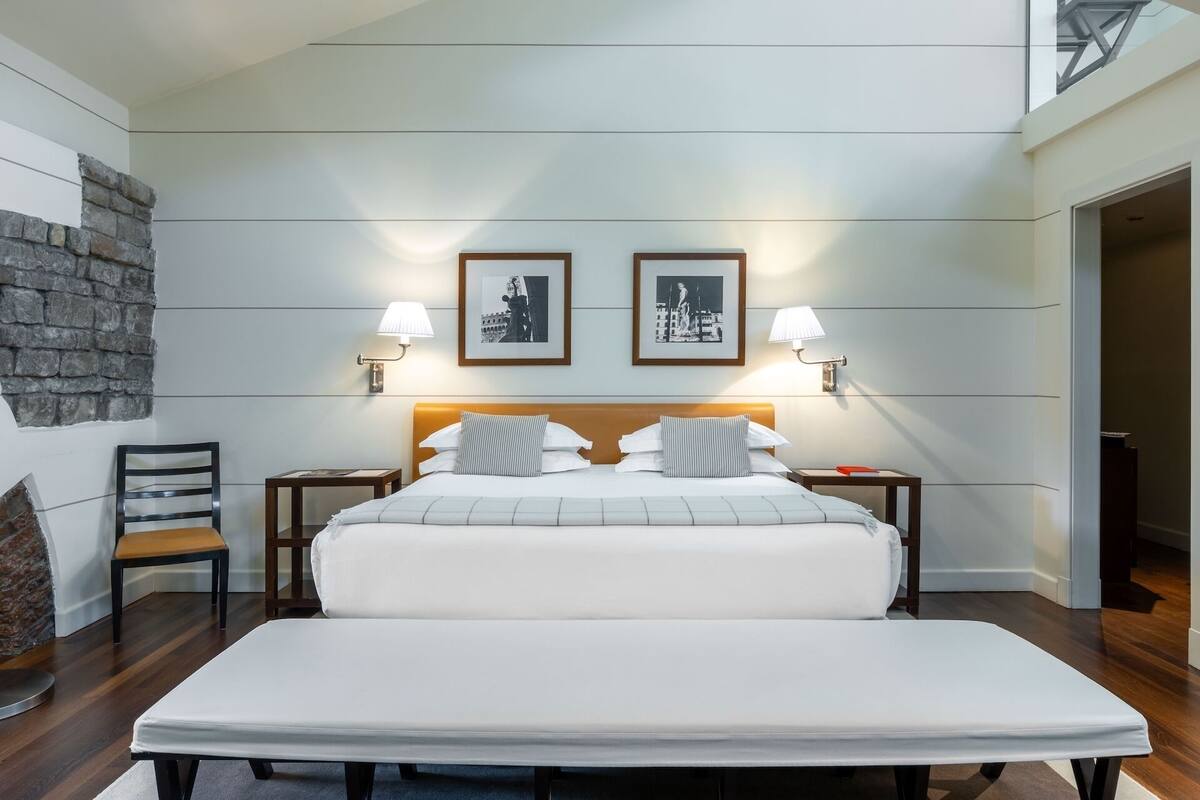 A hotel room with a large bed, two bedside tables with lamps, a chair, a stone wall, and two framed pictures on the wall.