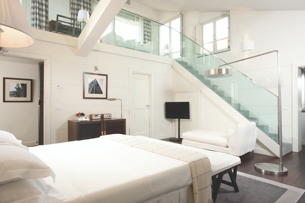 A modern hotel room with a large bed, a desk, a chair, a flat-screen TV, a desk lamp, a glass staircase, and a view of the city.