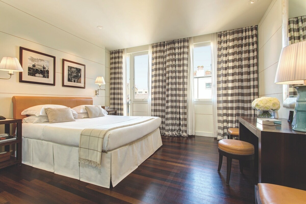 A hotel room with a bed, bedside tables, a desk, a chair, and curtains.