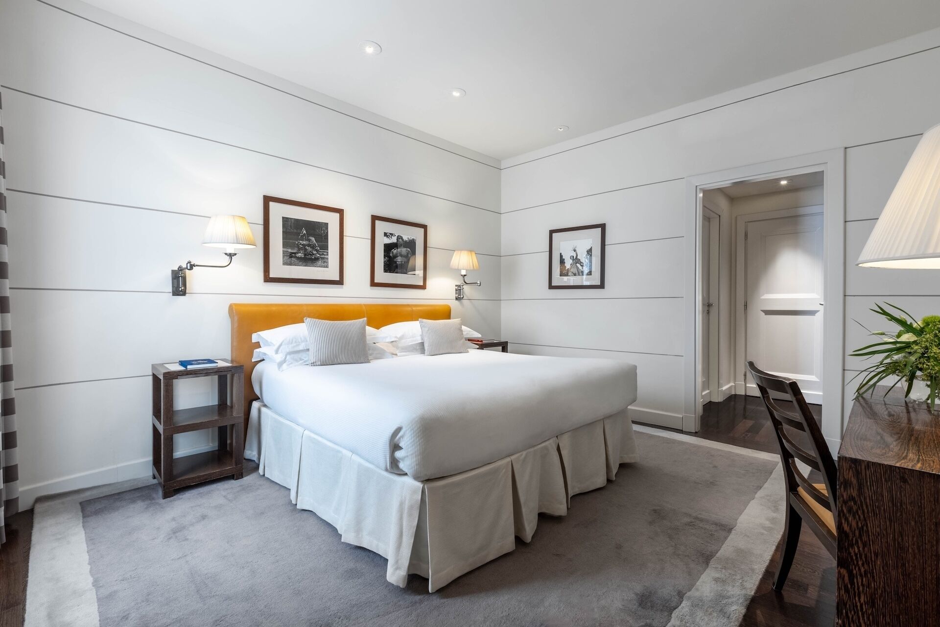 A hotel room with a large bed, bedside tables, wall-mounted lamps, framed pictures, and a door leading to another room.