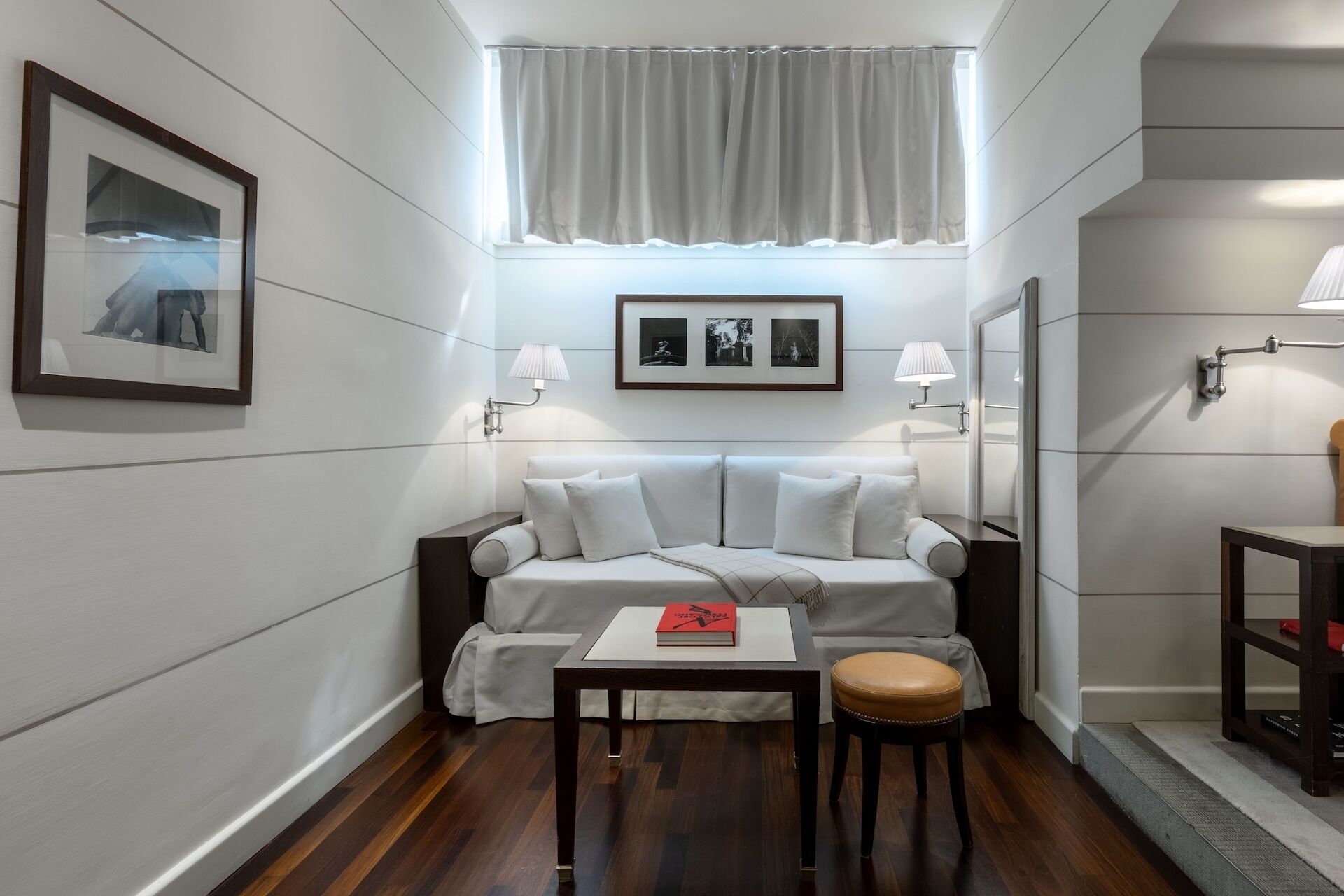A modern hotel room with a sofa, a small table, a chair, a framed picture, and a window with curtains.