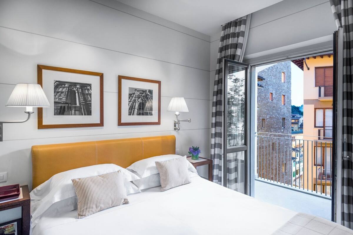 A hotel room with a bed, two framed pictures, a nightstand, a lamp, and a view of buildings through a window.