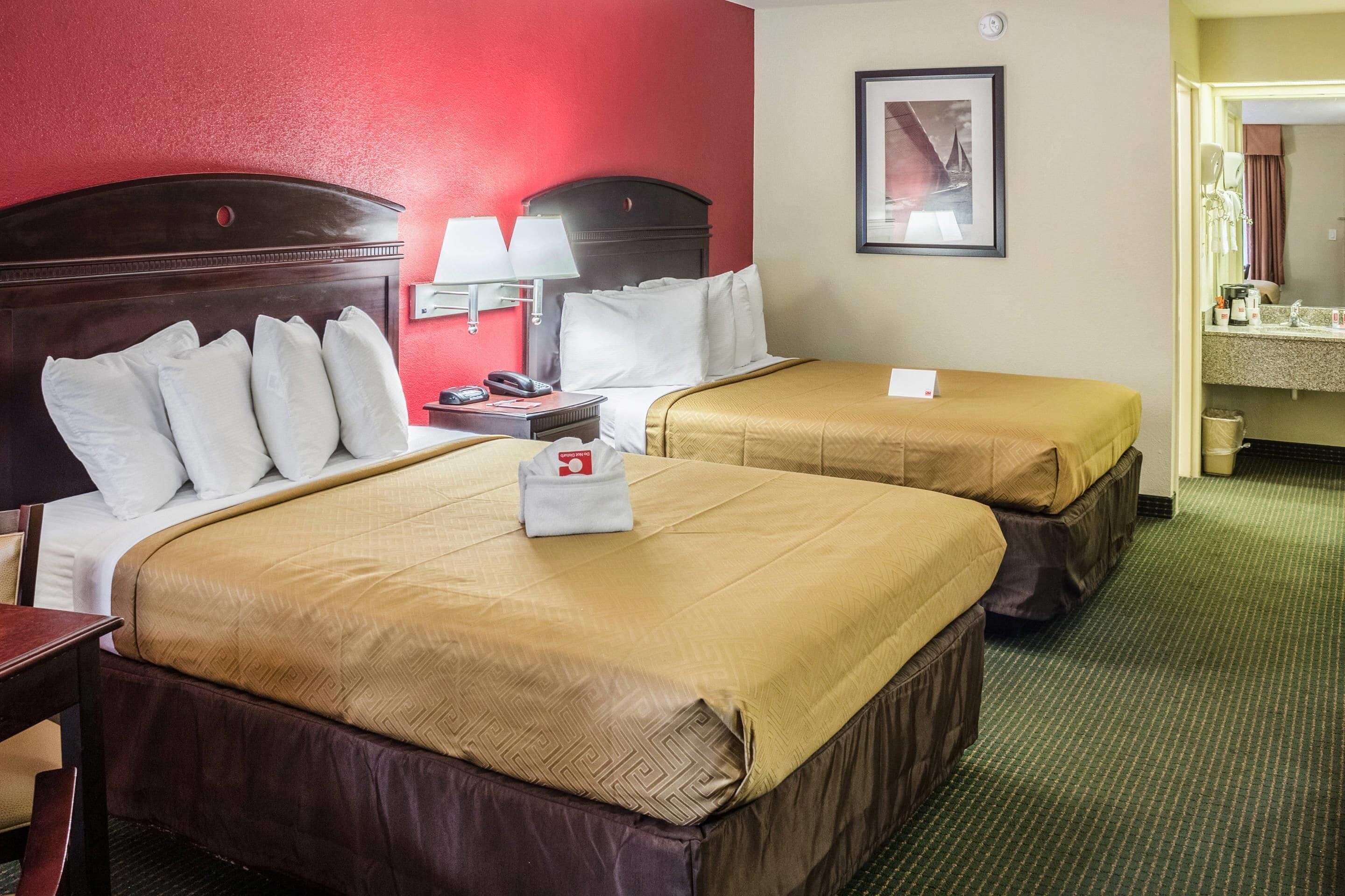 A hotel room with two beds, a nightstand, a lamp, a framed picture on the wall, and a bathroom visible through an open door.
