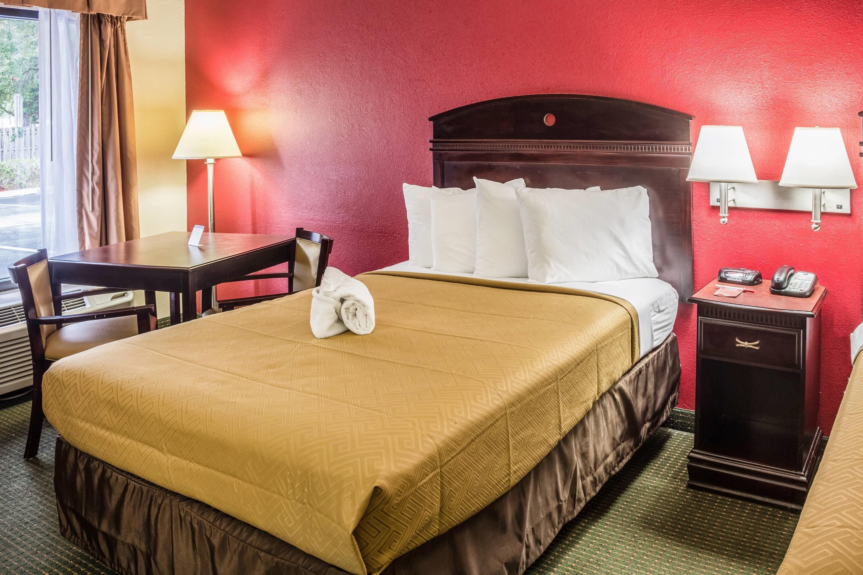 A hotel room with a bed, a desk with a lamp, a chair, a nightstand with a phone, and a window with curtains.