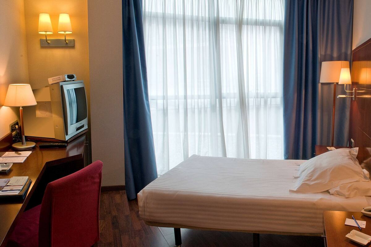 Une chambre d’hôtel avec un lit, un bureau avec une lampe, une chaise et une fenêtre avec des rideaux.