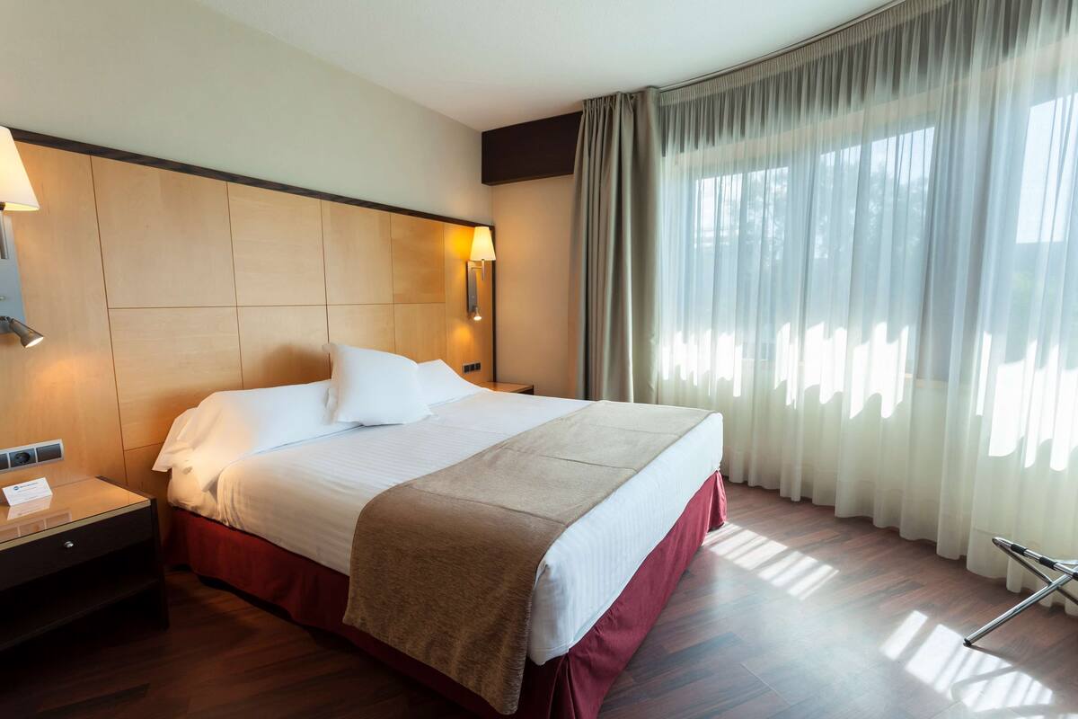 Une chambre d’hôtel avec un grand lit, une table de chevet, une lampe et une fenêtre avec des rideaux.