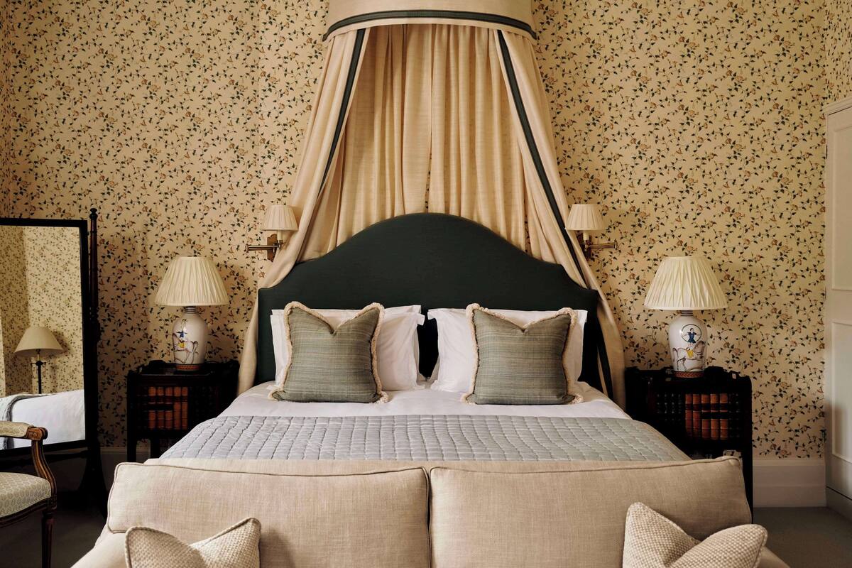 A bedroom with a four-poster bed, floral wallpaper, two bedside tables with lamps, and a chair.