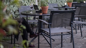 Un café en plein air, avec des tables et des chaises, entouré d’une végétation luxuriante et bordé d’un bâtiment à l’architecture classique.