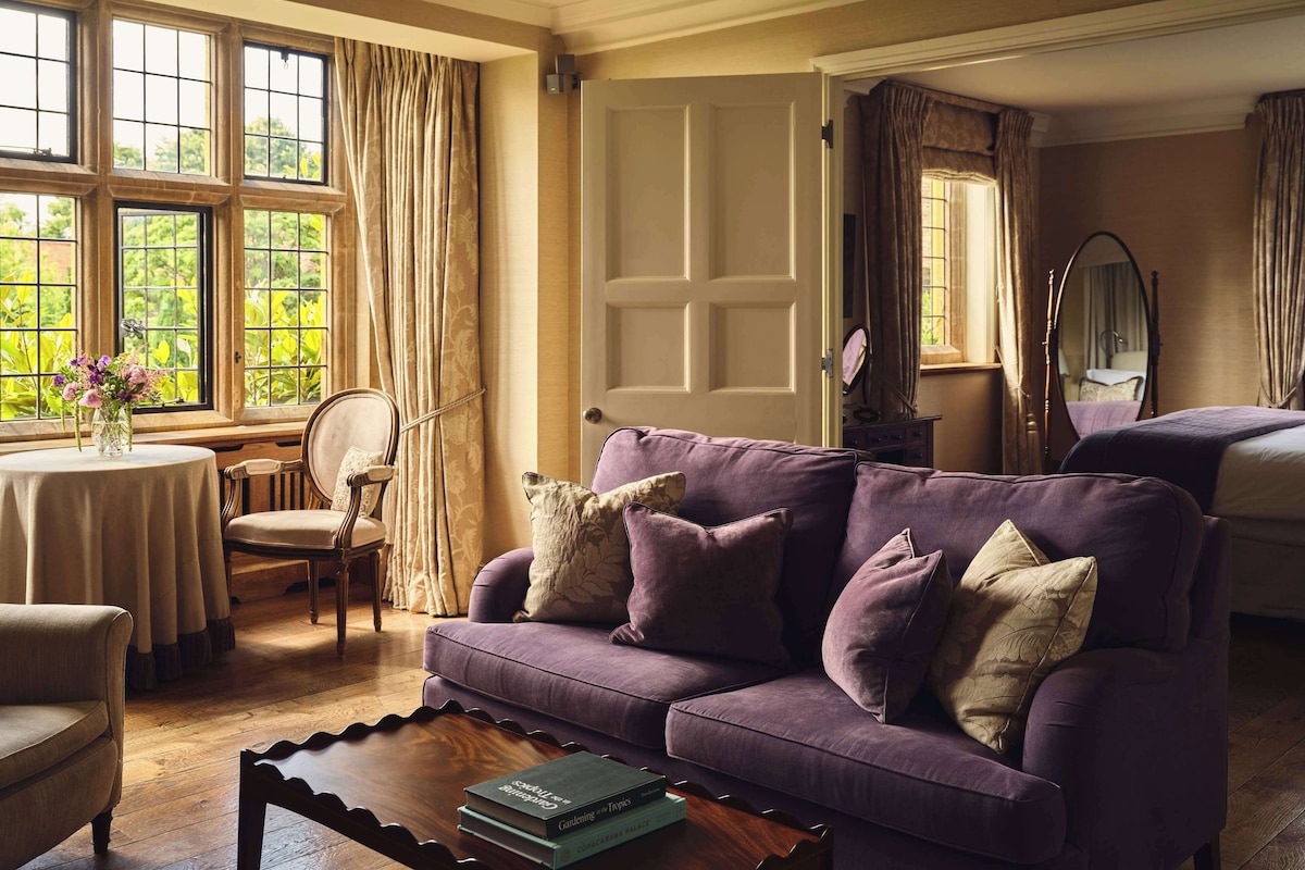 Une pièce avec un canapé violet, une table ronde ornée d’un vase de fleurs, une table basse en bois et un lit avec un miroir.