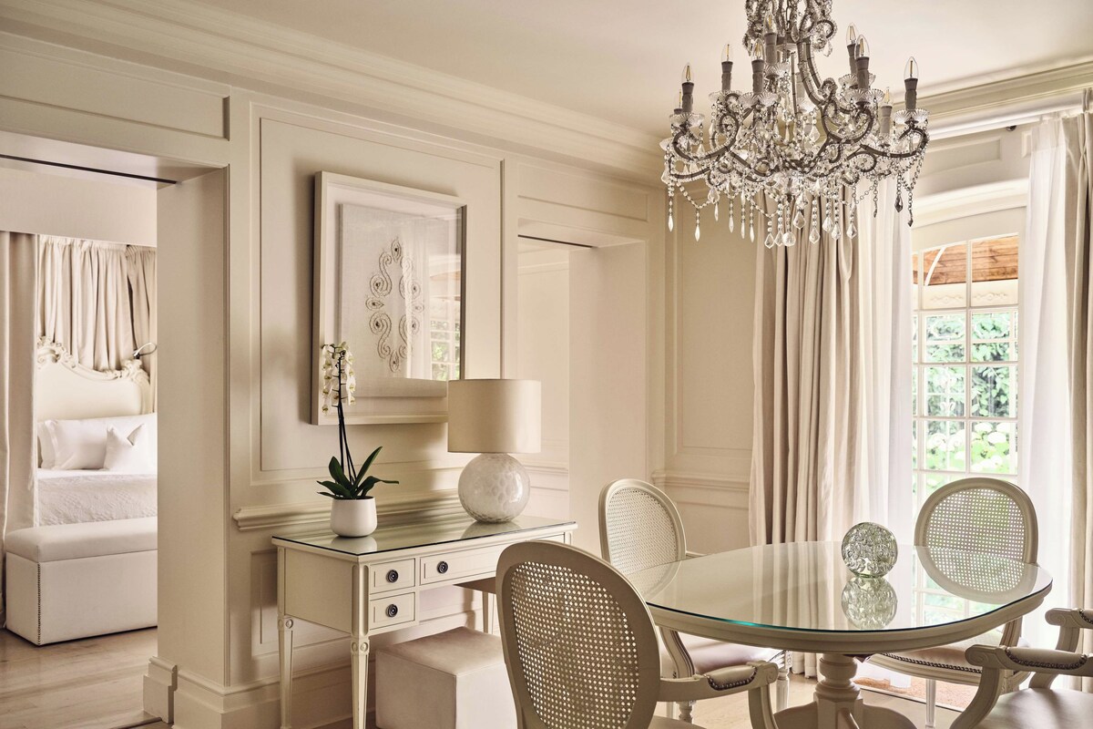 Uma sala de jantar com mesa de vidro, cadeiras brancas, um lustre, uma planta em vaso e uma obra de arte emoldurada na parede.