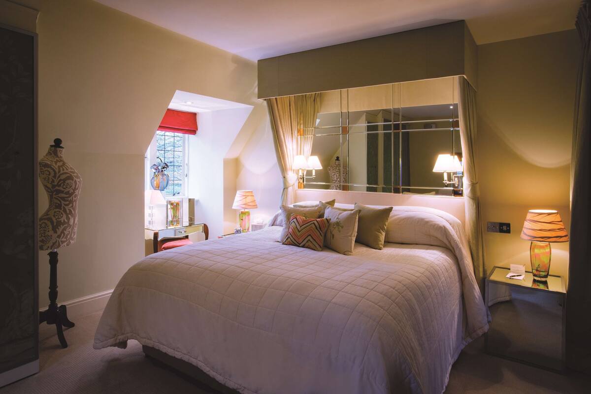 A neatly arranged bedroom with a large bed, a bedside table with a lamp, a mirror, and a window with curtains.