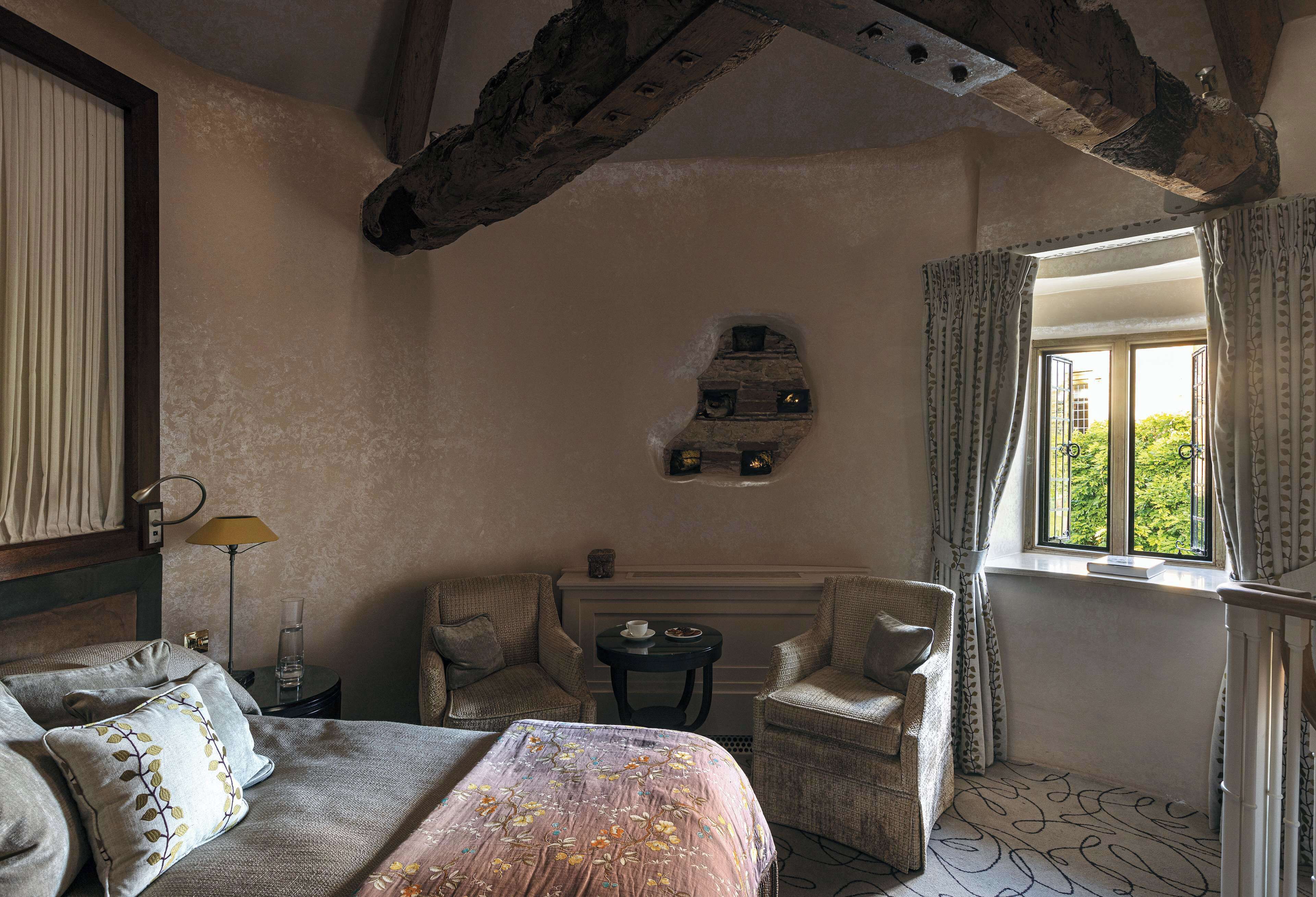 Une chambre comprenant un lit, deux fauteuils, une petite table, une fenêtre avec des rideaux et un mur en pierre décoratif.