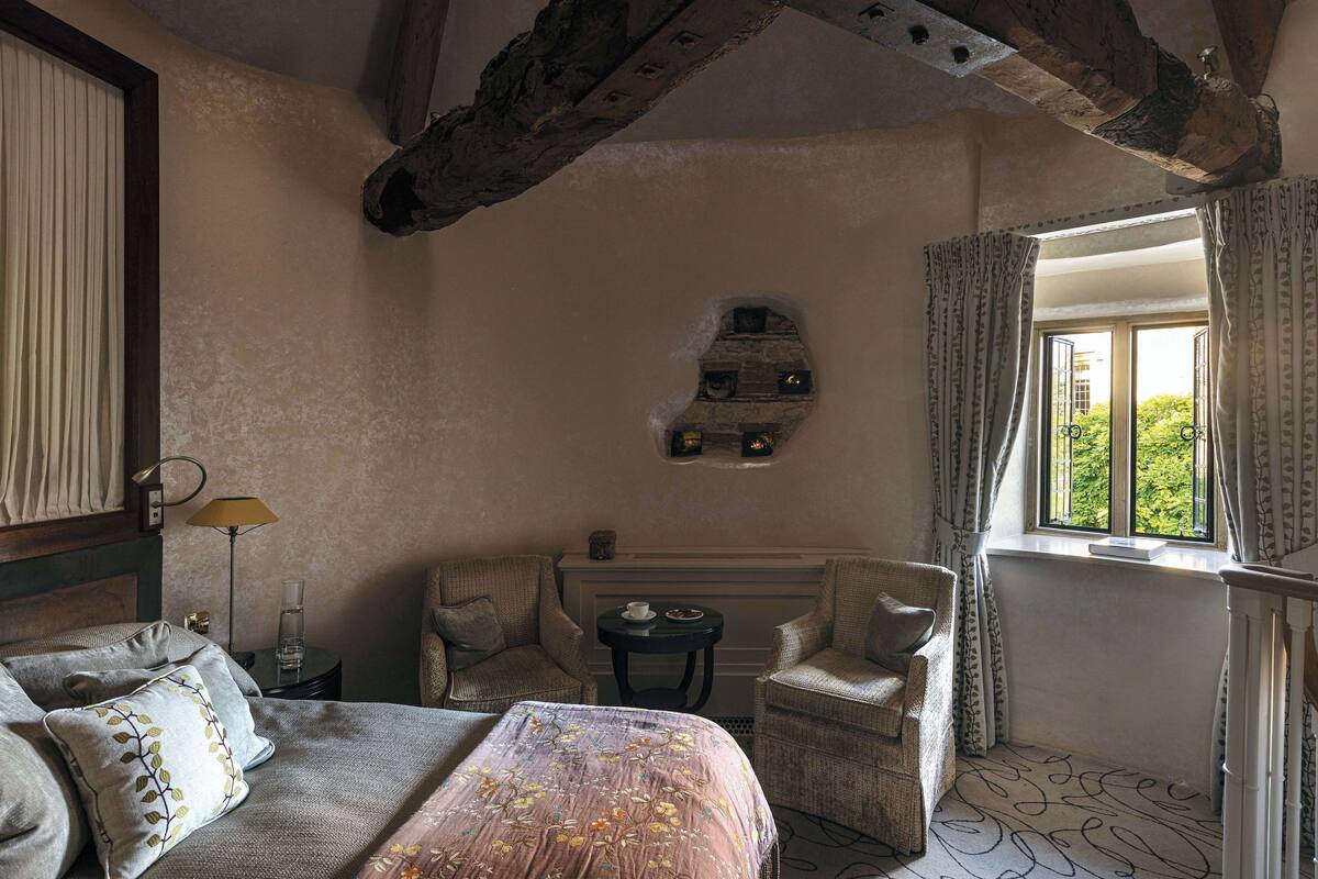 A bedroom with a bed, two armchairs, a small table, a window with curtains, and a stone wall feature.
