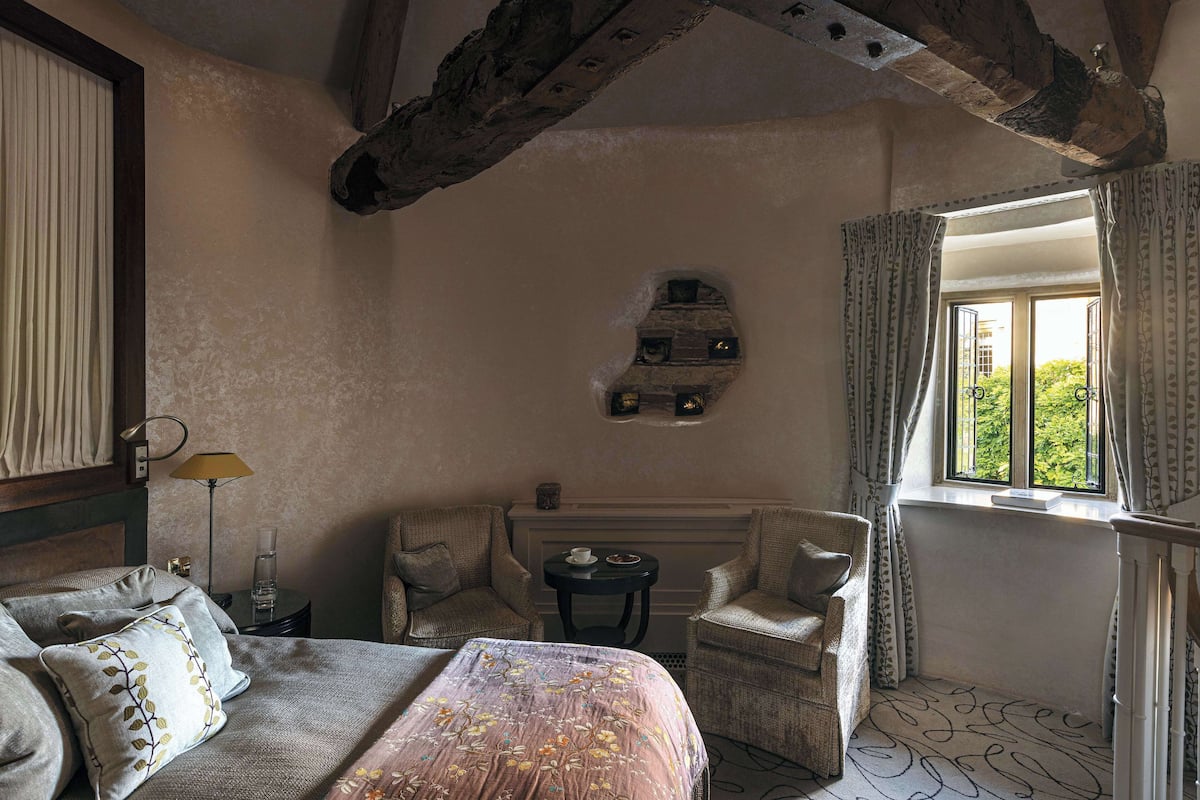 A bedroom with a bed, two armchairs, a small table, a window with curtains, and a stone wall feature.