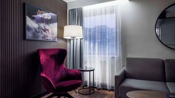 A modern hotel room with a dark wood panel wall, a red armchair, a grey sofa, a round coffee table, a floor lamp, and a large window with sheer curtains.