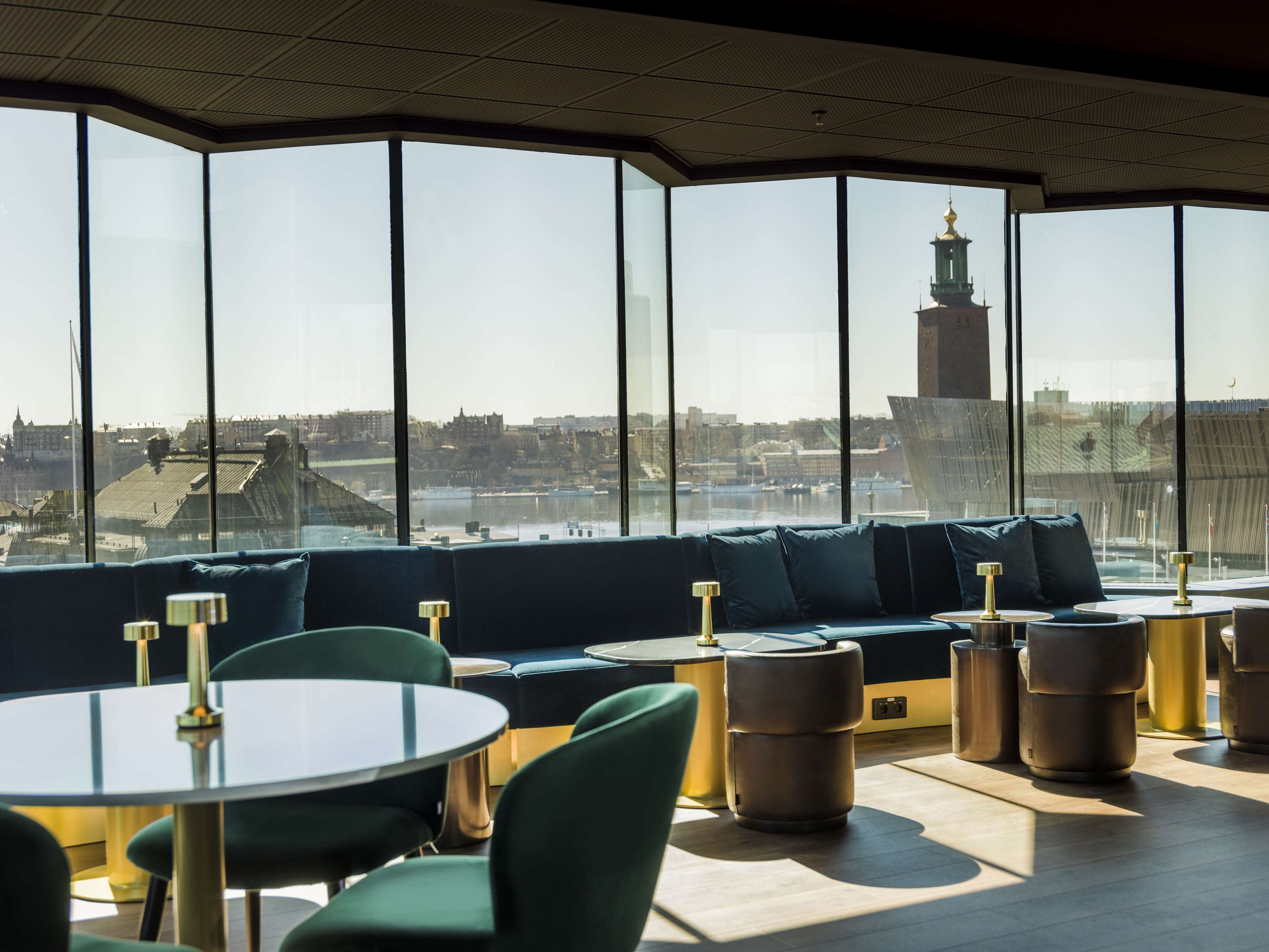 A modern lounge with a panoramic city view, featuring a curved seating area with round tables and chairs.