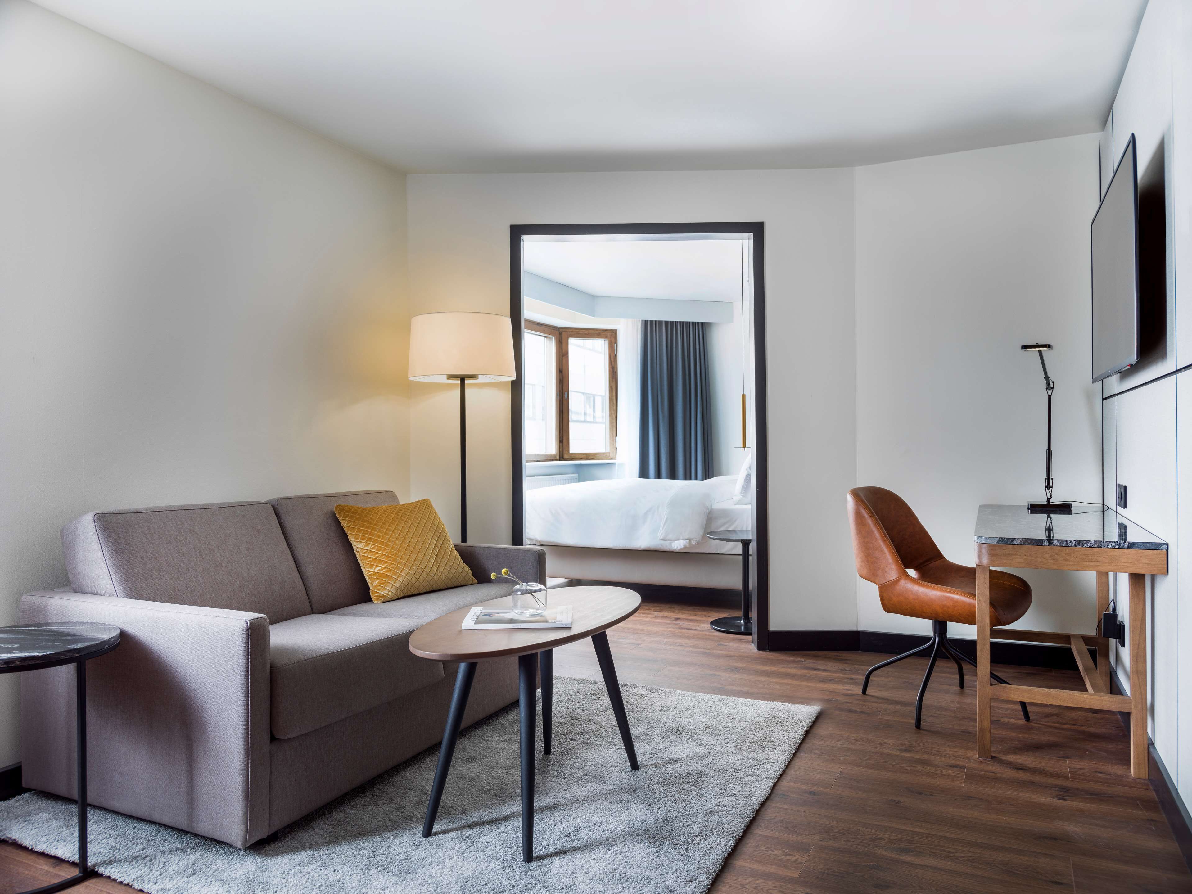 Una habitación de hotel moderna con sofá, una mesita redonda, un escritorio con silla, una lámpara y un televisor en la pared.