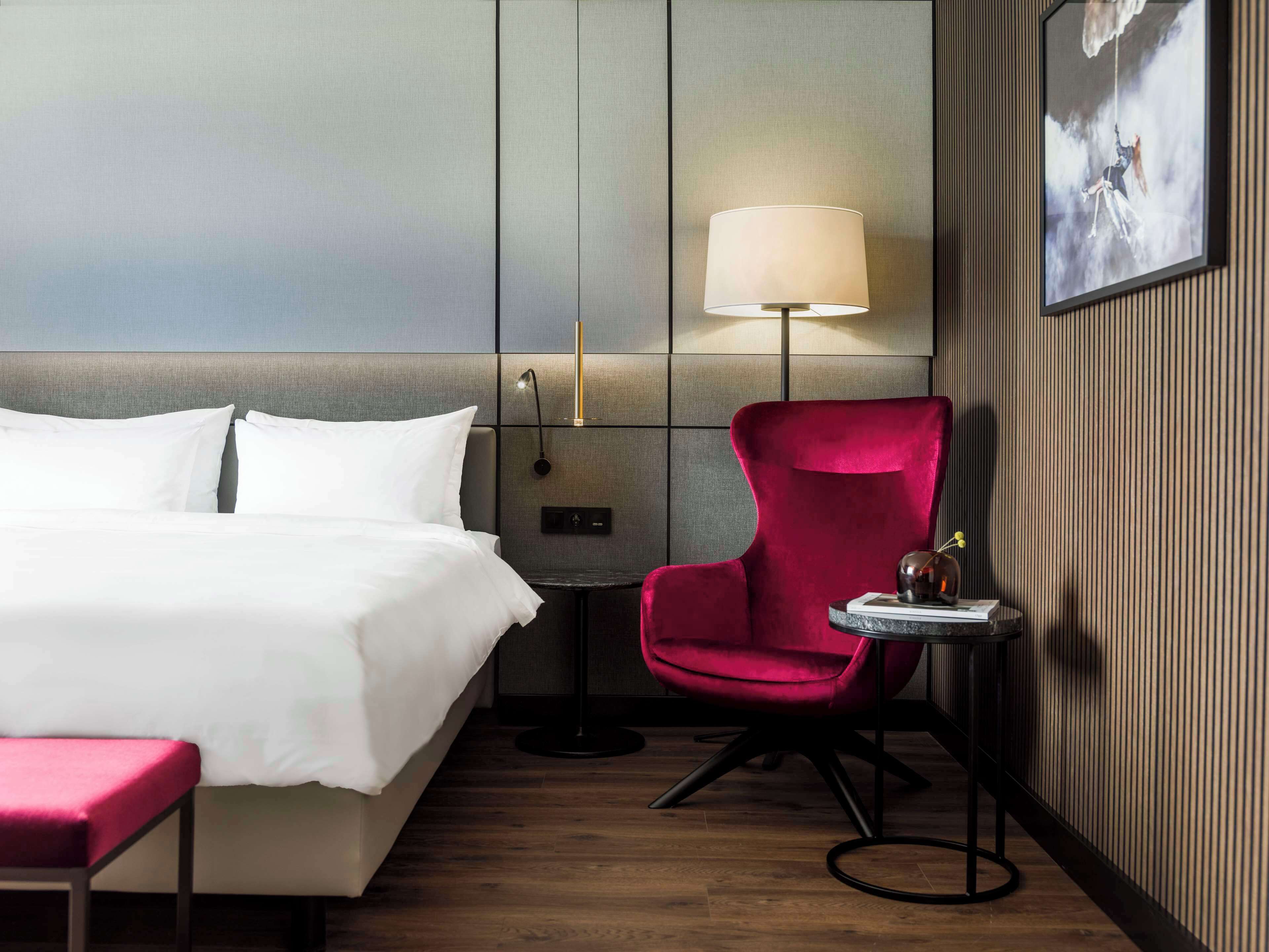A hotel room with a bed, a red chair, a small table with a lamp, and a framed picture on the wall.