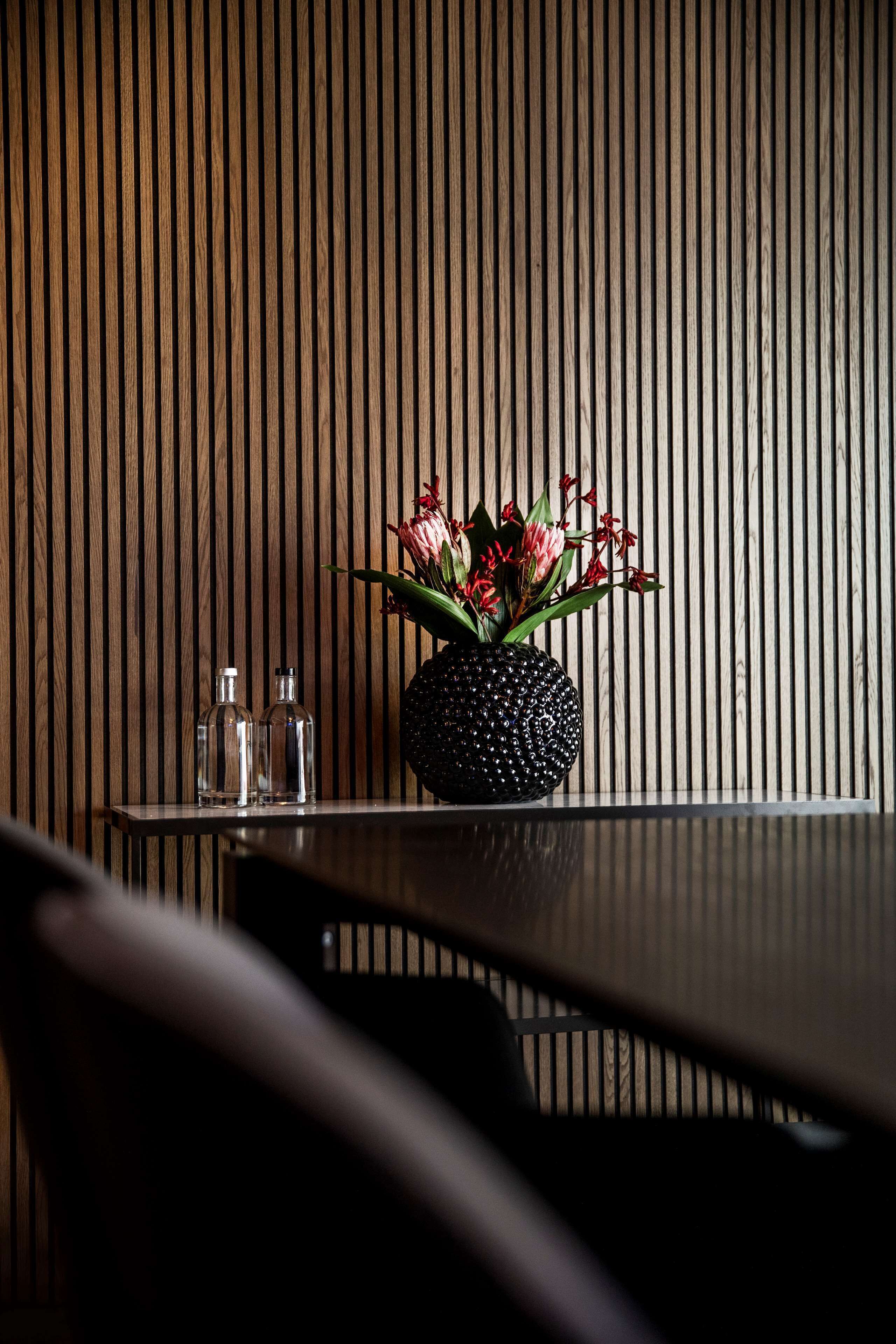 En vägg klädd med träribbor, en svart vas med blommor och två genomskinliga flaskor på ett bord.