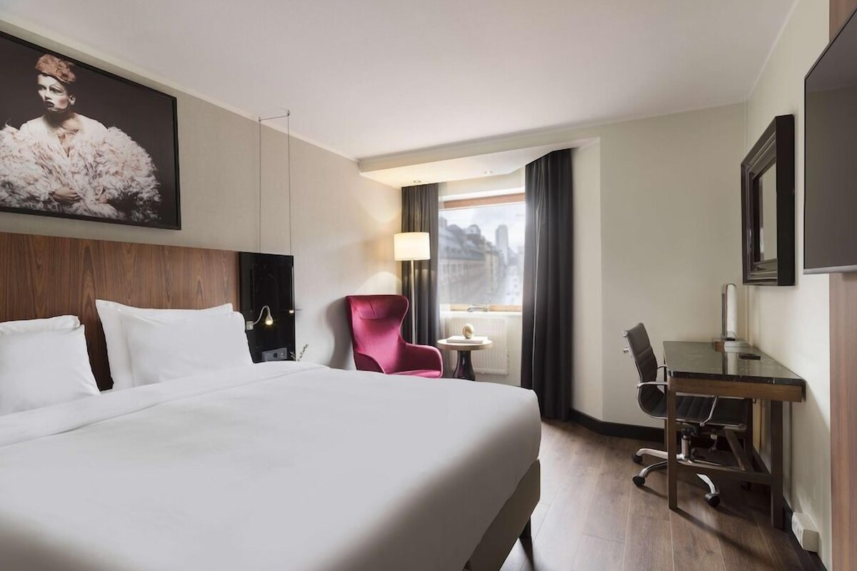 A hotel room with a large bed, a desk, a chair, a lamp, and a window with a view of buildings.
