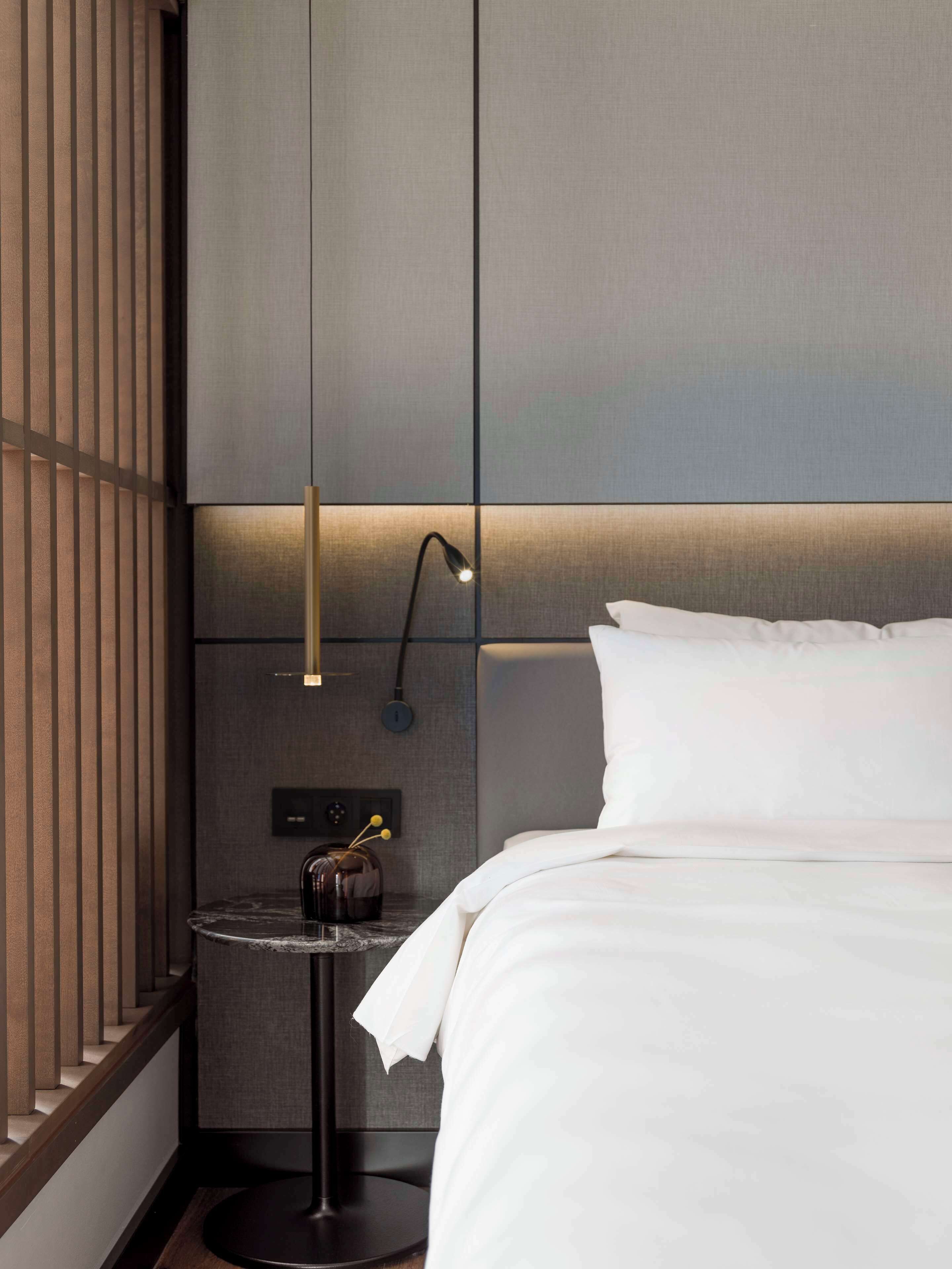A modern hotel room with a neatly made bed, a small round table with a vase, a wall-mounted lamp, and a textured wall panel.