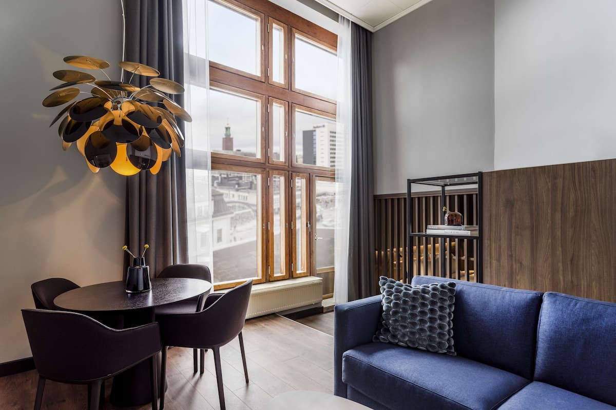 Un salon moderne avec un canapé bleu, une table à manger avec des chaises, un luminaire suspendu original et une grande fenêtre donnant sur des immeubles.